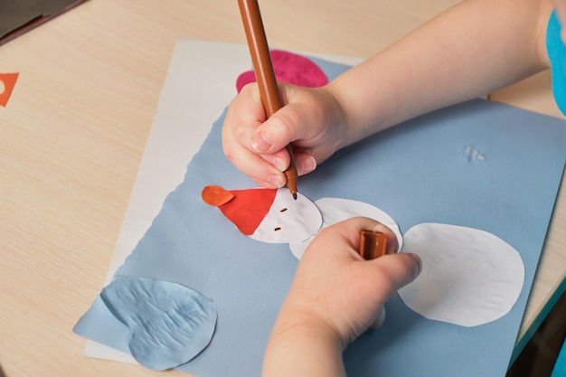 Ein Vorschulmädchen in einem blauen T-Shirt macht eine Applikation, während sie am Tisch sitzt, ein Papierschneemann, der auf farbigem Papier gezogen wird, Kinderkreativität im Winter, atopische Dermatitis an den Händen eines Kindes