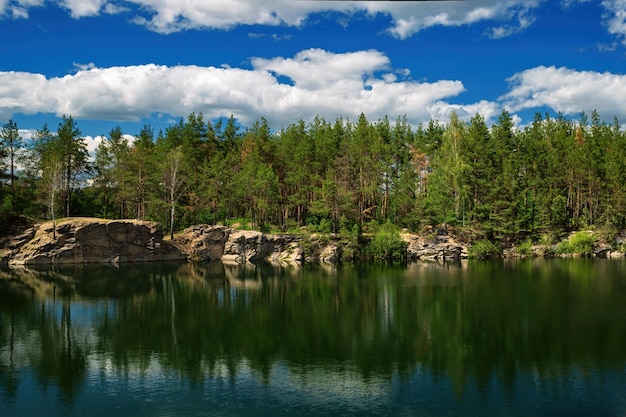 Ein Waldsee mit felsigen Ufern und Nadelbäumen mit blauem Himmel und weißen Wolken, die sich im Wasser spiegeln