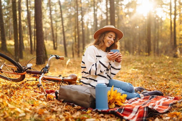 Ein Wanderer sitzt auf einer Picknickdecke und trinkt ein heißes Getränk im Herbstwald. Eine Frau ruht sich in der Natur aus.
