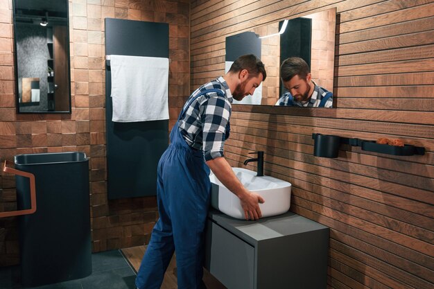 Foto ein waschbecken halten und bewegen. klempner in blauer uniform ist im badezimmer bei der arbeit