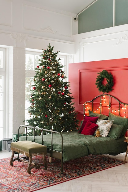 Ein weihnachtlich dekoriertes Schlafzimmer mit einem Weihnachtsbaum und einem Kranz an der Wand Ein Doppelbett in einem weihnachtlichen Interieur auf dem Hintergrund einer roten Wand
