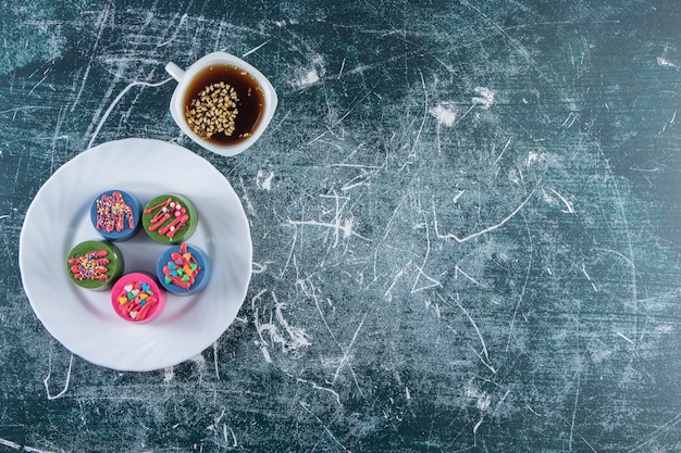 Ein weißer Teller mit bunten Cupcakes mit Streuseln und einer Tasse schwarzen Tee.