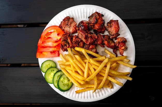 Ein weißer Teller mit einem Fleischgericht und Gemüse auf einem schwarzen Holztisch Gurken- und Tomatenscheiben neben gebratenen Fleischpommes Frites auf einem weißen Einwegteller