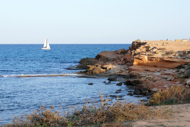 Foto ein weißes segelboot segelt auf dem meer gegen eine oberfläche aus küstenfelsen mit einer klippe.