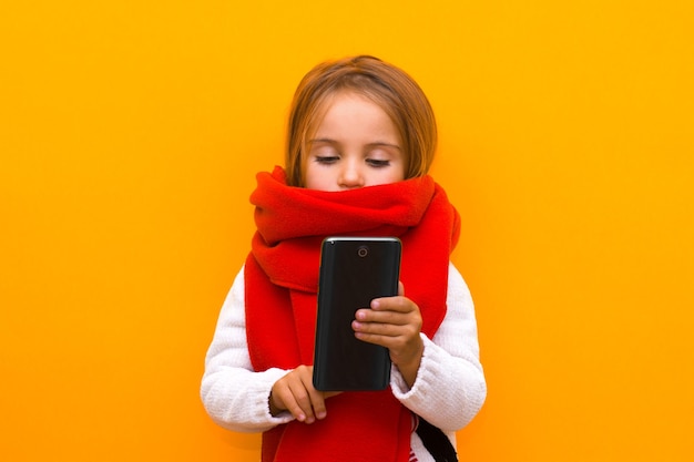 Ein Winterkind, das auf den Bildschirm eines Mobiltelefons schaut, wählt Geschenke auf einem isolierten gelben Hintergrund aus