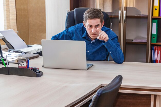 Ein wütender Chef, der mit einem Laptop an einem Tisch sitzt, zeigt mit dem Finger auf einen Untergebenen. Ein strenger Verweis an den Untergebenen. Büroangestellter. Abteilungsleiter.