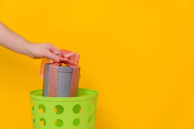 Ein wunderschön verpacktes Geschenk wird in einen grünen Mülleimer geworfen, gelber Hintergrund, Seitenansicht, Kopierraum