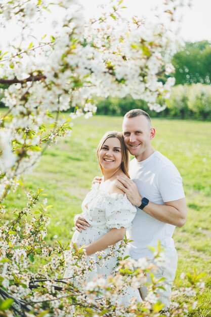 Ein wunderschönes junges Paar erwartet eine Schwangerschaft an einem romantischen Ort, einem im Frühling blühenden Apfelgarten. Fröhliches, fröhliches Paar, das sich beim Spaziergang im Garten amüsiert. Ein Mann hält die Hand einer Frau