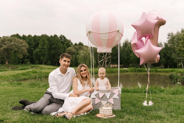 Ein wunderschönes kleines Mädchen mit Mama und Papa in einem Korb mit Ballon und Kuchen feiert ihren ersten Geburtstag