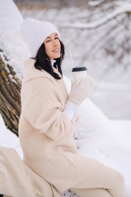 Ein wunderschönes Mädchen mit beiger Strickjacke und weißem Hut, das in einem verschneiten Winterwald in der Nähe eines Sees Tee trinkt