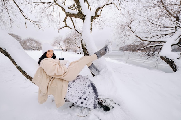 Ein wunderschönes Mädchen mit beiger Strickjacke und weißem Hut, das in einem verschneiten Winterwald in der Nähe eines Sees Tee trinkt