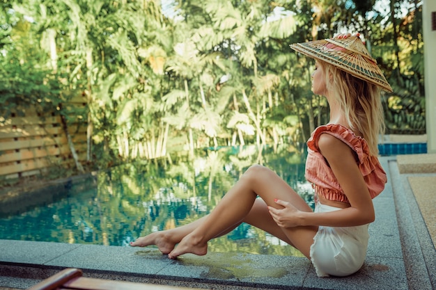 Ein zerbrechliches blondes Mädchen sitzt in einem asiatischen Hut nahe dem Pool. Ruhe in heißen Ländern