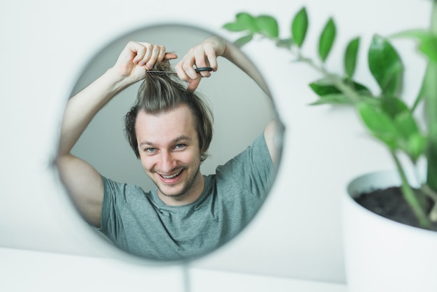 Ein zotteliger Mann in einem grauen T-Shirt mit einer Schere in den Händen versucht, sich die Haare zu schneiden, während er in den Spiegel schaut.