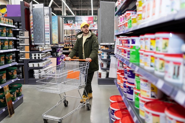 Foto ein zufriedener männlicher kunde läuft mit einem einkaufswagen in der hand zwischen den warenreihen in einem baumarkt