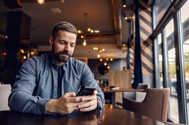 Ein zufriedener Mann sitzt am Tisch im Café und scrollt am Telefon