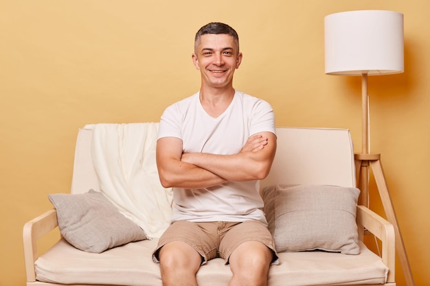 Ein zuversichtlicher, positiver, lächelnder brünetter junger Mann in einem bequemen weißen T-Shirt sitzt auf dem Sofa gegen die beigefarbene Wand und posiert mit gekreuzten Armen und schaut mit glücklichem Gesichtsausdruck in die Kamera