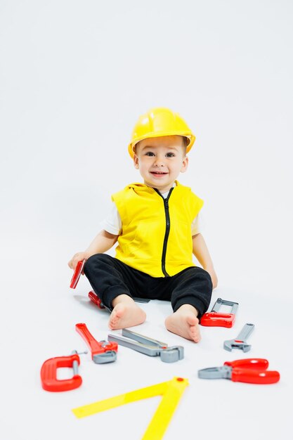 Ein zweijähriger Junge in einer gelben Weste sitzt und spielt mit Plastikbauwerkzeugen auf weißem Hintergrund. Spielzeug für kleine Kinder