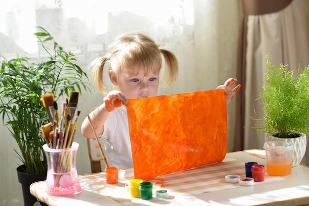 Ein zweijähriges Mädchen sitzt an einem Tisch und zeigt ein mit Farben bemaltes orangefarbenes Blatt. Taktile Entwicklung der Hände