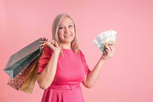 Eine 40-jährige erwachsene Frau mit Einkaufstüten und einem Fächer von Euro-Scheinen in der Hand auf rosa Hintergrund, Exemplar