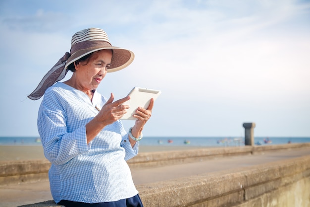 Eine ältere asiatische Frau spielt das Internet über ein Tablet, um sich zu entspannen und das Meer zu besuchen.