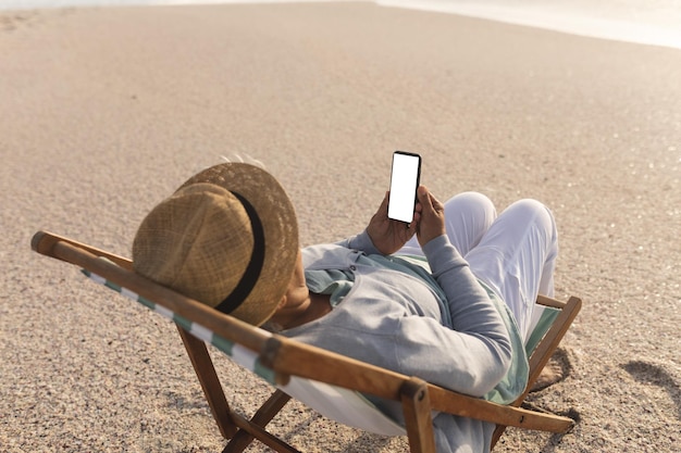 Eine ältere, biraciale Frau im Ruhestand benutzt ein Mobiltelefon mit leerem Bildschirm und Kopierplatz auf einem Stuhl am Strand