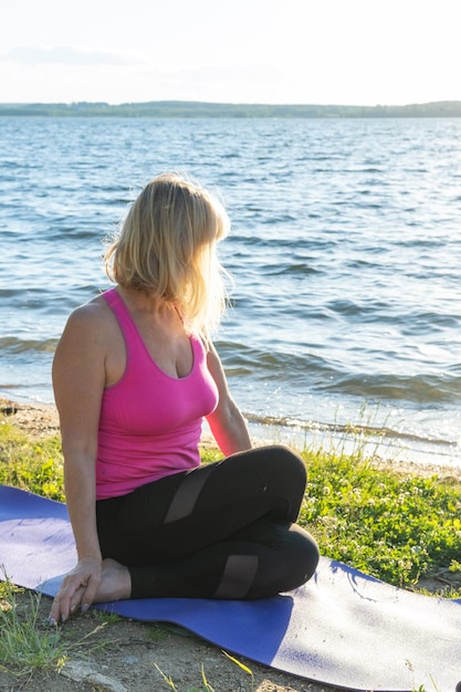 Eine ältere Frau, die im Sommer Yoga am Ufer eines Sees praktiziert Meditationssport im Alter Yoga