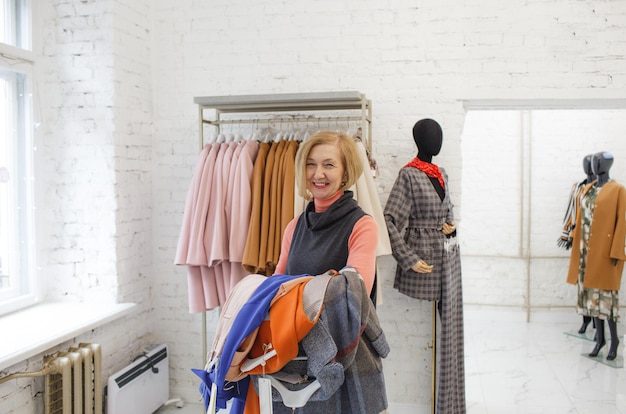 Eine ältere Frau hält einen Kleiderhaufen in ihren Händen und bereitet sich darauf vor, in einem Bekleidungsgeschäft in die Umkleidekabine zu gehen Einkaufen und Einkaufen