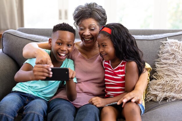 Eine ältere Frau mit gemischter Abstammung und ihre Enkelkinder genießen ihre gemeinsame Zeit zu Hause, sitzen auf einer Couch, umarmen sich, lächeln, benutzen ein Smartphone und machen ein Selfie