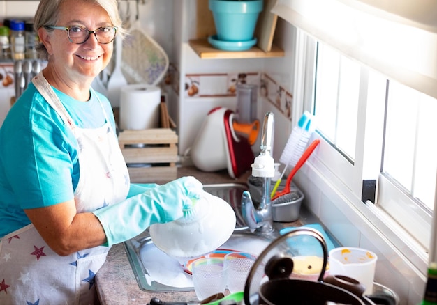 Eine ältere Frau mit grauen Haaren, die Schürze und Handschuhe beim Abwasch trägt Ecke der Küche Großmutter bei der Arbeit Helles Licht aus dem Fenster