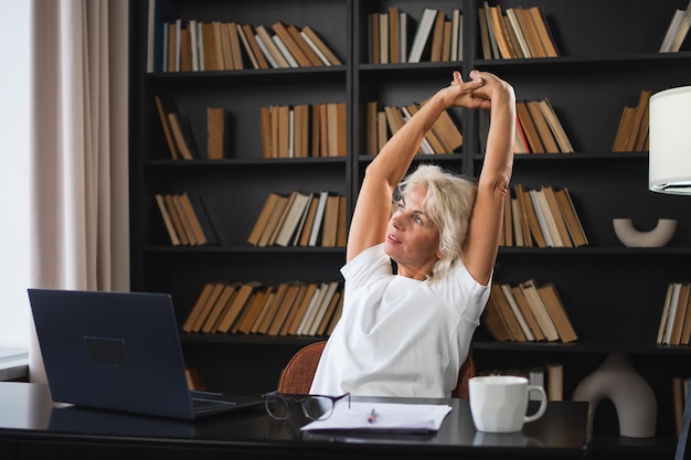 Eine ältere Frau mittleren Alters, die sich entspannt, Hände und Körper streckt und eine Pause von der Arbeit am Laptop zu Hause macht