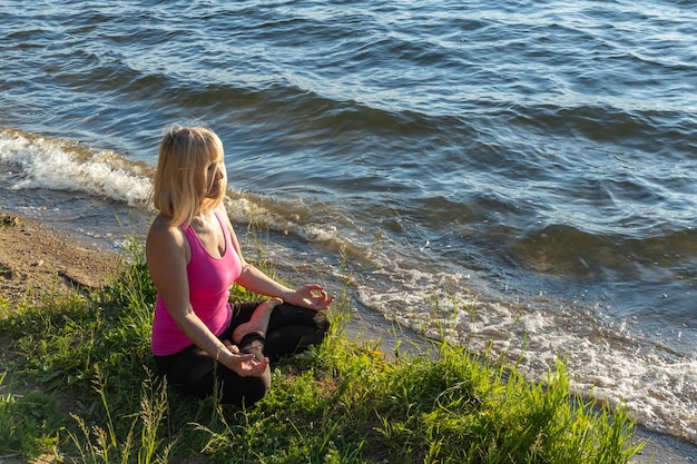 Eine ältere Frau sitzt im Lotussitz auf einer Sportmatte am Meeresufer Meditationssport