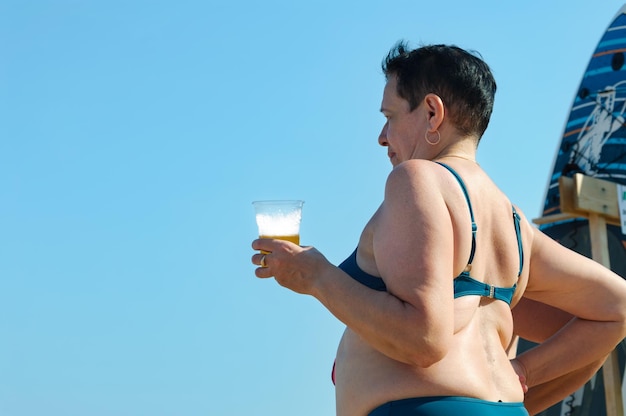 Eine ältere Frau steht mit einem Glas Bier am Strand