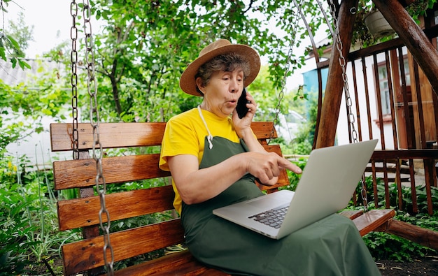 Eine ältere Gärtnerin mit Hut sieht sich etwas auf einem Laptop an und führt im Freien einen Videoanruf im Hof durch