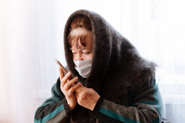 Eine ältere Großmutter mit einer chirurgischen medizinischen Maske und einem Taschentuch mit einem Telefon in den Händen