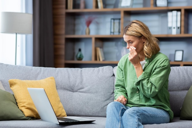 Eine ältere schöne Frau sitzt zu Hause auf dem Sofa mit einem Laptop weint und wischt ihre Tränen mit einer Serviette ab