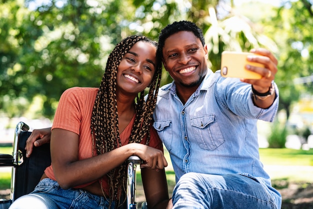 Eine Afroamerikanerin im Rollstuhl, die ein Selfie mit ihrem Freund nimmt, während sie einen Tag im Park zusammen genießt