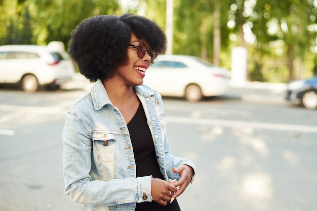 Eine Afroamerikanerin steht mit Sonnenbrille auf einer Stadtstraße und lacht.