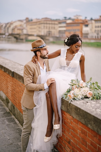 Eine afroamerikanische Braut sitzt auf einer Mauer und ein kaukasischer Bräutigam umarmt sie. Der Damm des Arno mit Blick auf die Stadt und die Brücken. Interracial Hochzeitspaar