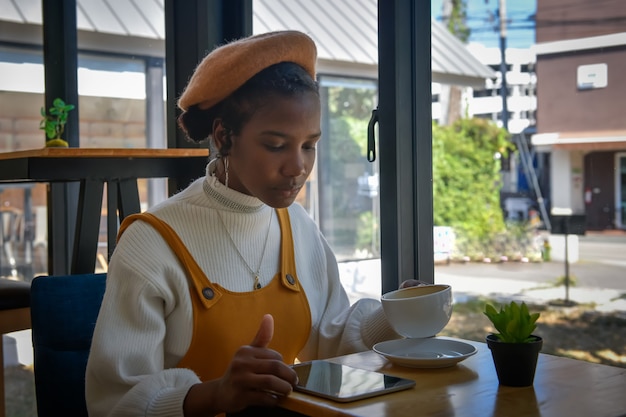 Eine afroamerikanische Geschäftsfrau verwendet ein Tablet, um mit Kunden in einem Café zu chatten und eine Kaffeetasse in der Hand zu haben.