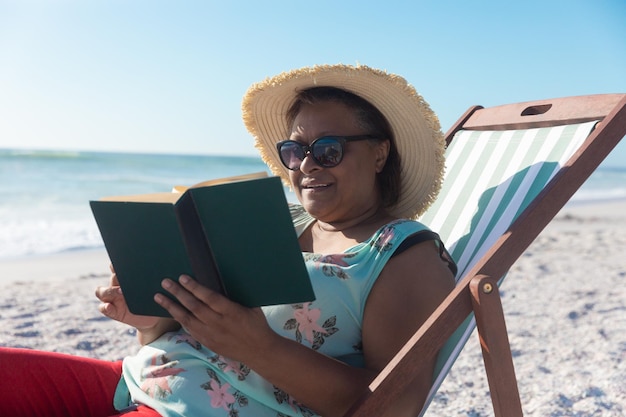 Eine afroamerikanische Rentnerin im Ruhestand liest ein Buch, während sie auf einem Klappstuhl am Strand sitzt