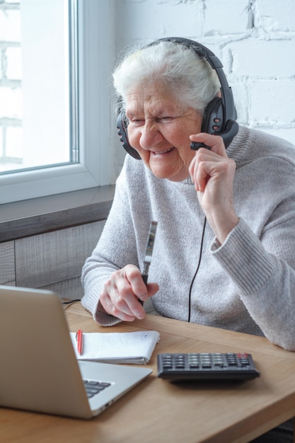 Eine alte Frau sitzt an einem Tisch vor einem Laptop mit Kopfhörern und schreibt in ein Notizbuch.