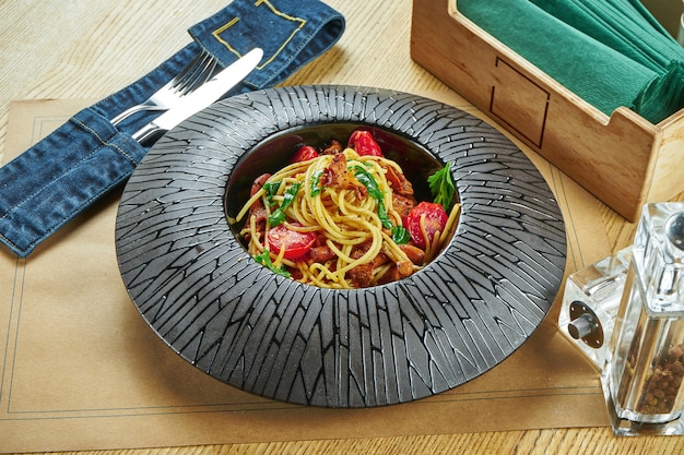 Eine appetitliche Portion hausgemachter italienischer Pasta (Spaghetti) mit Waldpilzen (Pfifferlingen), Kirschtomaten und Ruccola in einem schwarzen Teller auf einem Holztisch. Tischdekoration im Restaurant