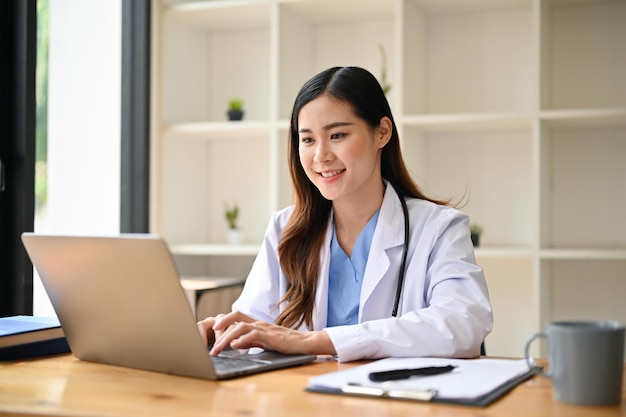 Eine asiatische Ärztin, die in ihrem Krankenhausbüro an ihrer medizinischen Forschung auf einem Laptop arbeitet