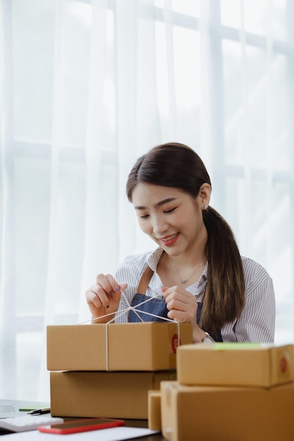 Eine asiatische Frau, die ein Paket an die Kiste eines Kunden bindet, besitzt einen Online-Shop, den sie verpackt und über ein privates Transportunternehmen versendet. Online-Verkaufs- und Online-Shopping-Konzepte