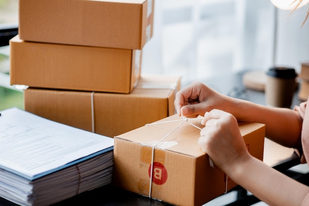 Eine asiatische Frau, die ein Paket an die Kiste eines Kunden bindet, besitzt einen Online-Shop, den sie verpackt und über ein privates Transportunternehmen versendet. Online-Verkaufs- und Online-Shopping-Konzepte