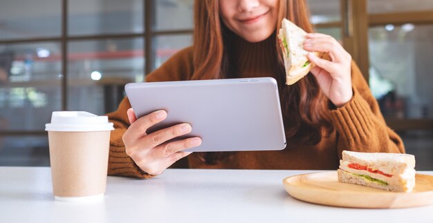 Eine asiatische Frau, die ein Vollkornsandwich hält und isst, während sie einen Tablet-PC verwendet