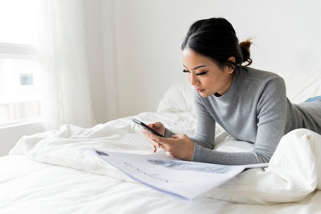 Eine asiatische Frau, die einen Handy auf einem Bett überprüft