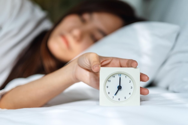 Eine asiatische Frau, die morgens einen Wecker ausschaltet, während sie auf einem weißen, gemütlichen Bett schläft