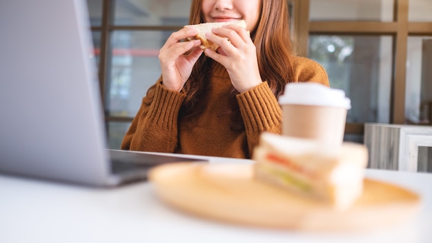 Eine asiatische Frau, die Vollkornbrot hält und isst, während sie an einem Laptop arbeitet