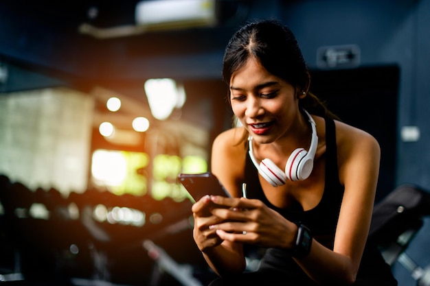 Eine asiatische Frau in Fitnesskleidung sitzt auf einem weißen Telefon und Kopfhörern und entspannt sich während des Trainings. Sie erzeugt Energie für Inspiration für sportliche Fitness